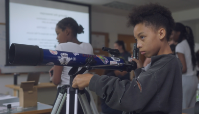 A young girl adjusts a large blue telescope in a classroom.